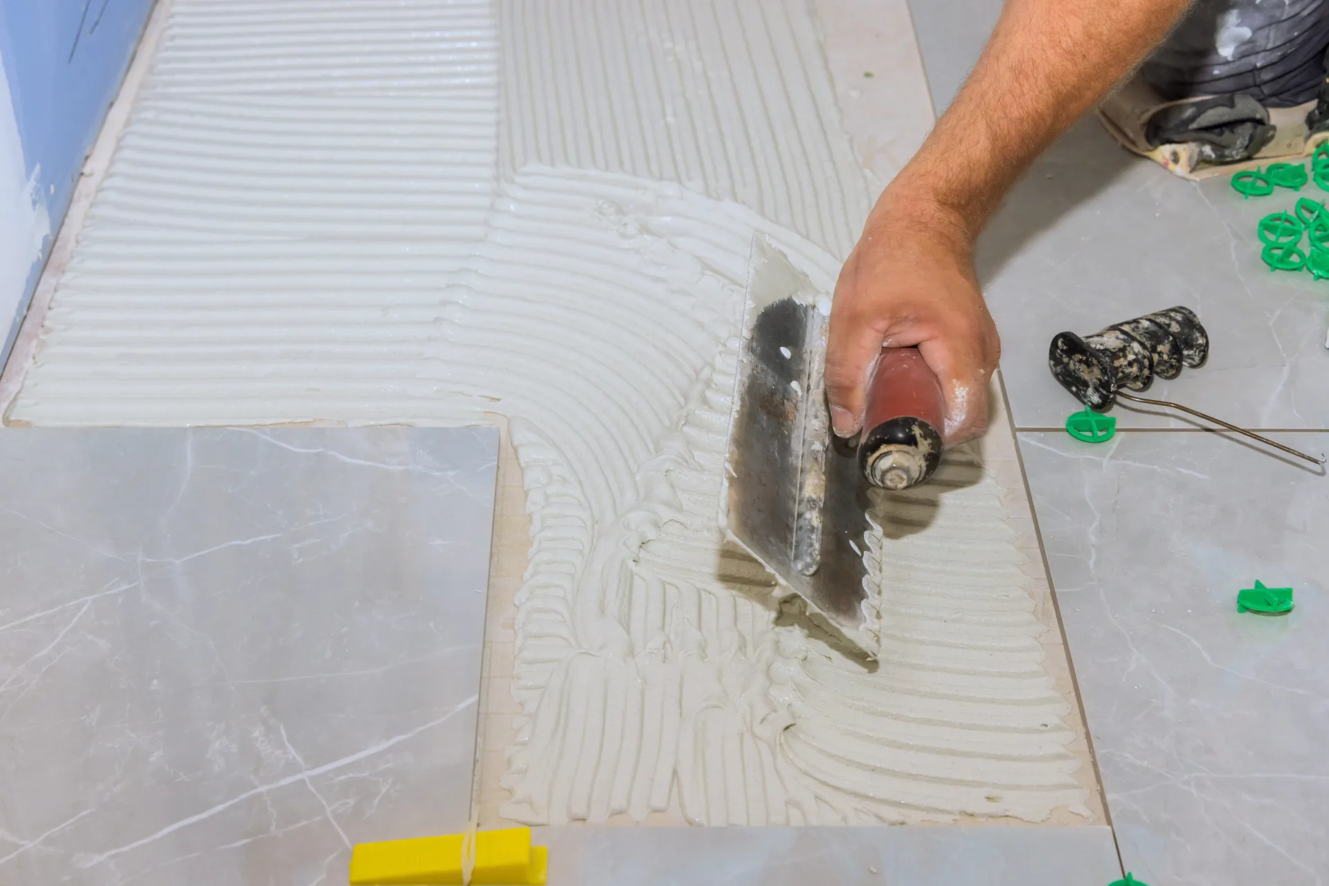 Notched trowel combing tile adhesive across a porcelain floor.