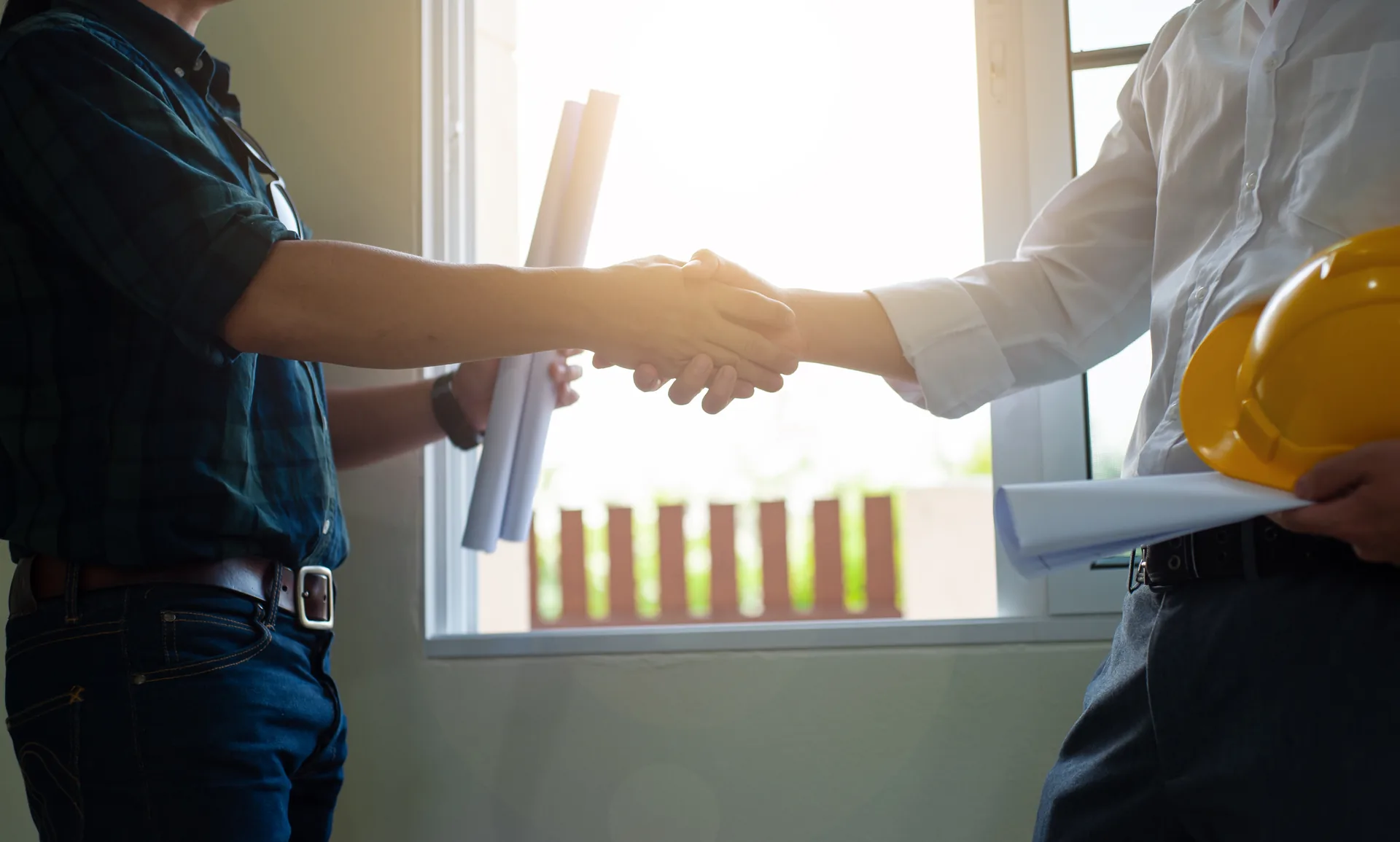 Two tradesmen shaking hands beside a window holding plans.